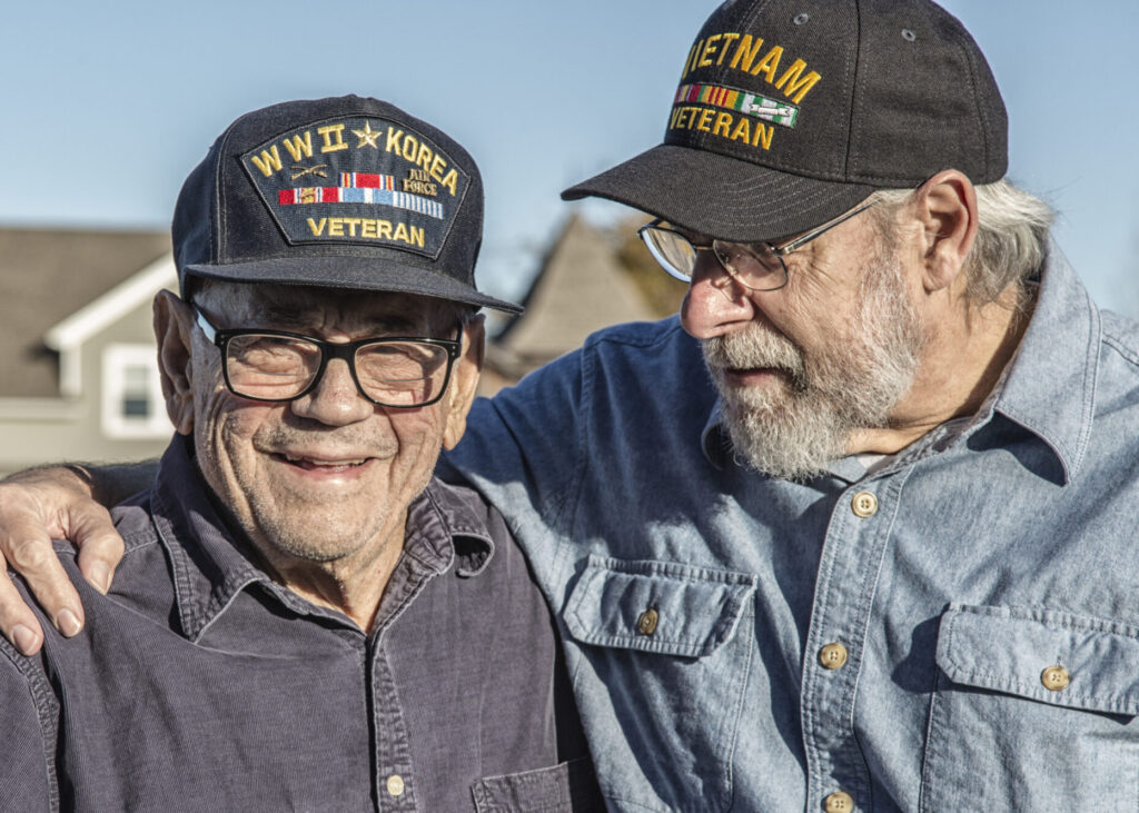 Two generations of real person senior adult men USA military veterans. On the left, a 93 year old United States Army World War II and Korean Conflict US Air Force military veteran is smiling at the camera while his son-in-law on the right - a 65 year old US Navy sailor Vietnam War military veteran - is teasing him - trying to distract him by telling jokes during the photo shoot. They are both wearing generic souvenir shop military veteran commemorative baseball style caps.